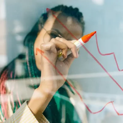 A Black woman wearing glasses is drawing a graph in red pen on a clear board.