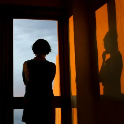 A person is standing with their arms crossed and their back to the camera, looking out a window at sunset.