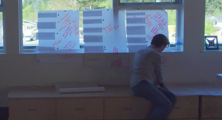 A person is sitting on a bench and taping up poster board to a window. The posters are covered in multi-colored writing.